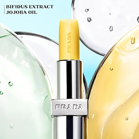 Image 5 - A yellow textured lip balm with PRADA embossed vertically on the stick is held in a silver tube featuring PRADA on its band, set against an abstract background of swirling green, clear, and golden liquids with bubbles, and the text BIFIDUS EXTRACT JOJOBA OIL is displayed in the top left.